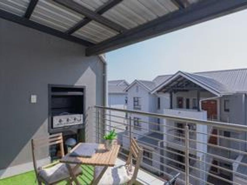 Covered balcony featuring a built-in braai, stainless steel balustrade, and artificial grass flooring, offering an elevated view of the surrounding apartment complex.