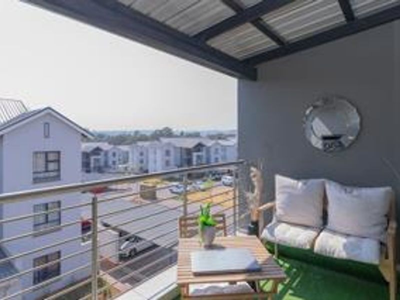 A covered balcony featuring a stainless steel balustrade and artificial grass flooring, offering an elevated view of the surrounding apartment complex.
