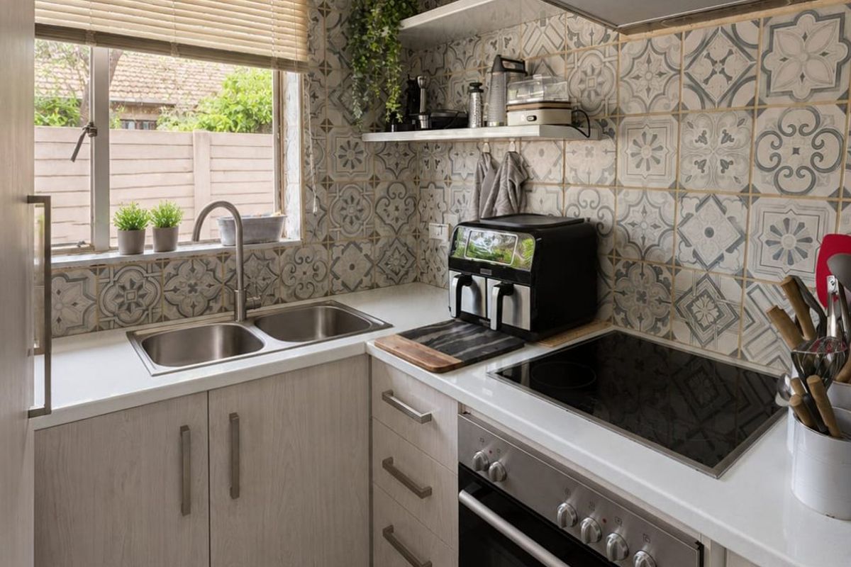 Compact, well-maintained kitchen featuring patterned wall tiles, modern cabinetry, stainless steel appliances, and a double bowl sink with a window view.