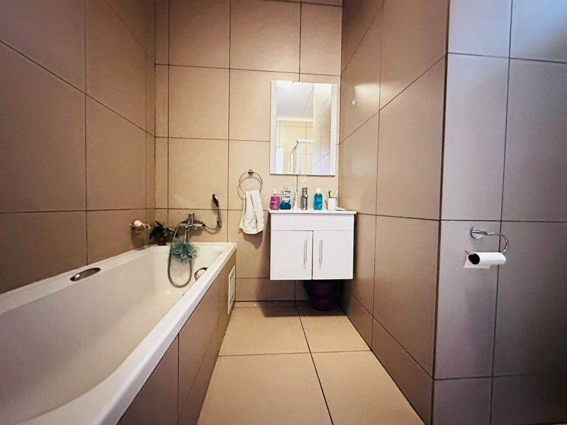 A functional bathroom featuring full-height wall tiling, a built-in bathtub with shower mixer, and a compact wall-mounted vanity unit.