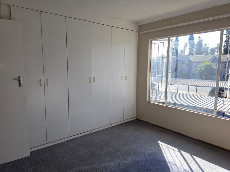 A bright, empty bedroom featuring extensive built-in cupboards, carpeted flooring, and large windows with security bars overlooking the complex grounds.
