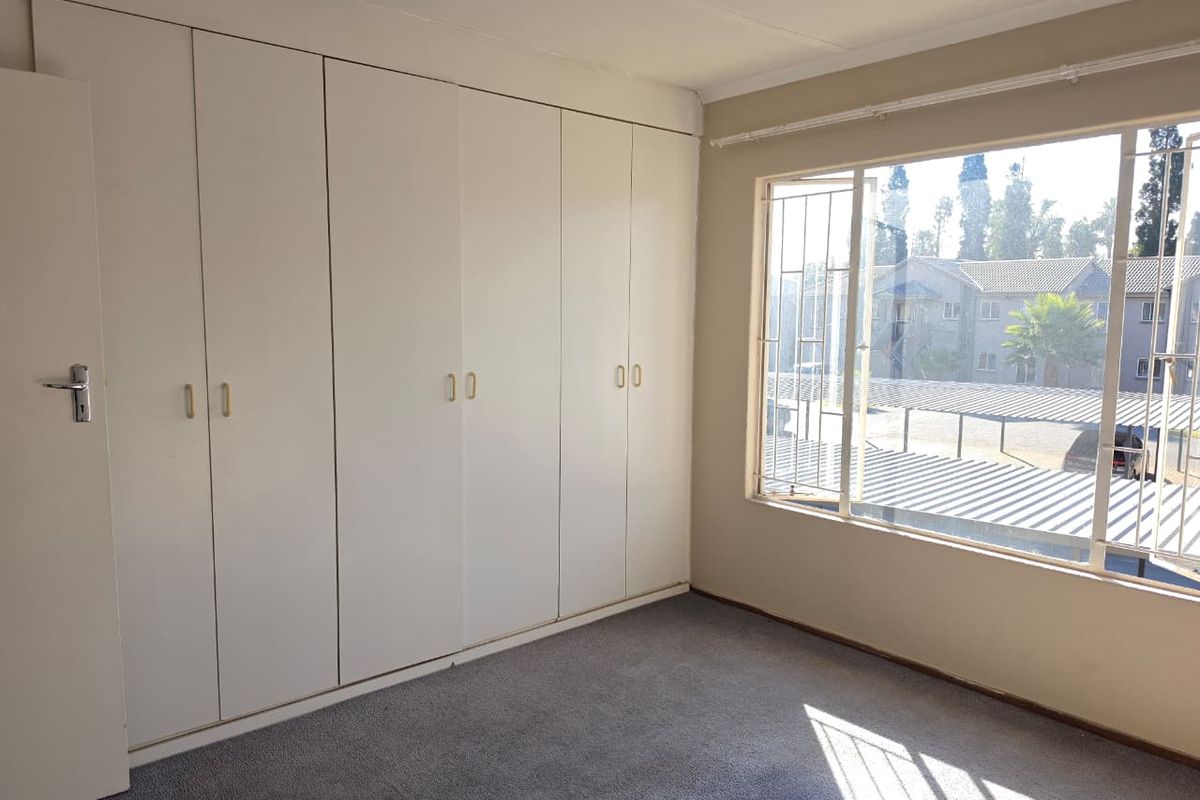 A bright, empty bedroom featuring extensive built-in cupboards, carpeted flooring, and large windows with security bars overlooking the complex.