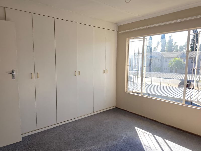 A bright, empty bedroom featuring extensive built-in cupboards, carpeted flooring, and large windows with security bars overlooking the complex.