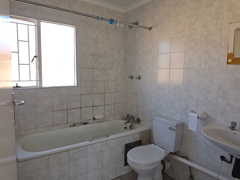 A dated bathroom featuring a bathtub, toilet, and pedestal basin with fully tiled walls and security bars on the window.