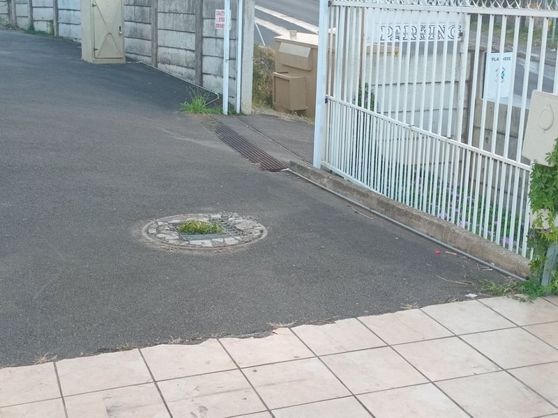 View of the apartment complex entrance featuring an asphalt driveway, metal security gate, and tiled patio area with some visible wear.