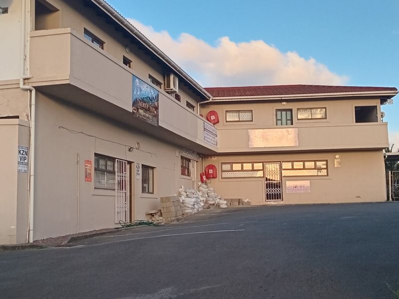 Front exterior view of a multi-level apartment building featuring an asphalt driveway, security fencing, and visible signage for commercial tenants.