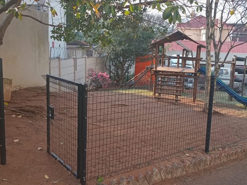 A fenced communal playground area featuring a metal gate, boundary wall, and basic play equipment situated adjacent to a paved driveway.