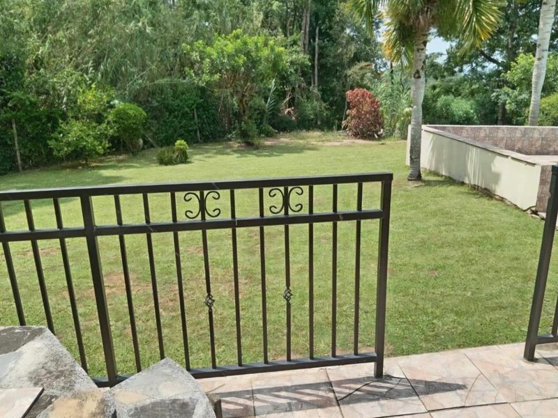 A view of a private garden featuring a tiled patio area with a wrought iron railing overlooking a well-maintained lawn and mature trees.