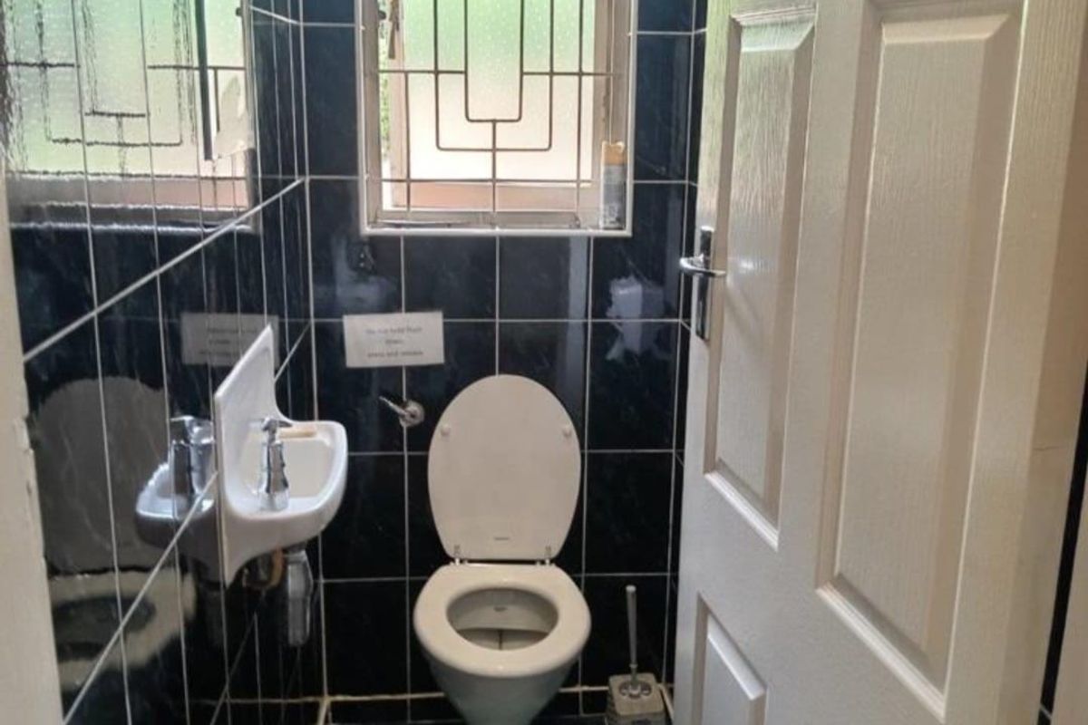 A compact guest bathroom featuring dark tiled walls and flooring, a wall-mounted basin, and a standard toilet with security bars on the window.