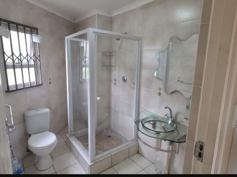 A functional bathroom featuring a glass vessel sink, a corner shower enclosure, and neutral wall tiling with security bars on the window.