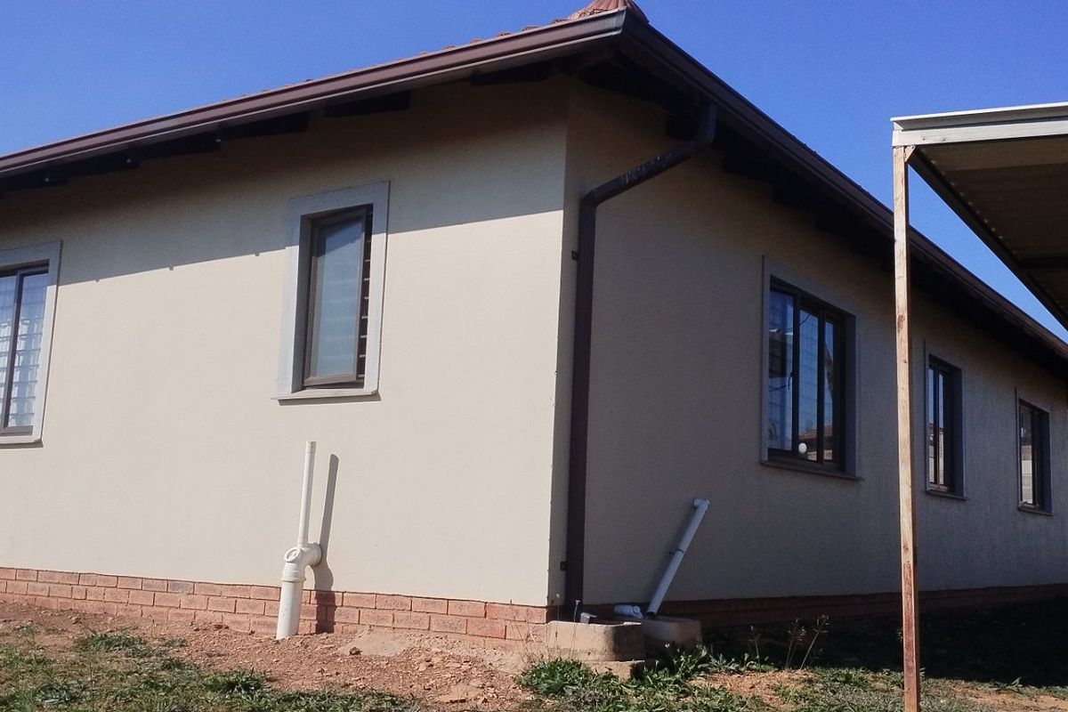 Rear view of a single-story house featuring a tiled roof, aluminium window frames, brick plinth, and an attached metal carport structure.