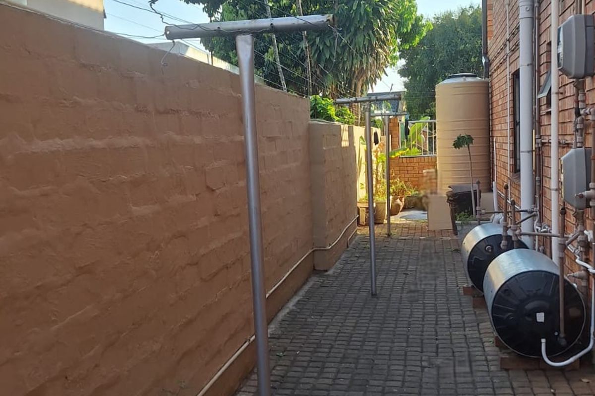 A side exterior area featuring a paved walkway, brick boundary wall, external electric geysers, and a fixed washing line.