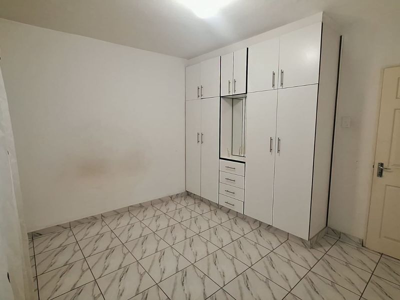 A clean, empty bedroom featuring white built-in cupboards with integrated drawers and vanity mirror, complemented by modern marble-look tiled flooring.