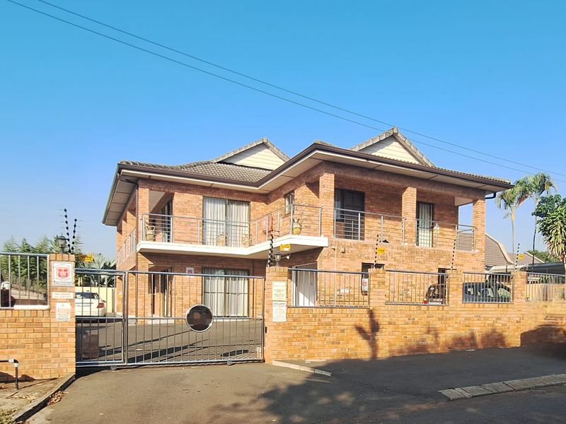 A double-story face brick apartment building featuring a tiled roof, upper-level balconies with metal railings, and a secure gated entrance with electric fencing.