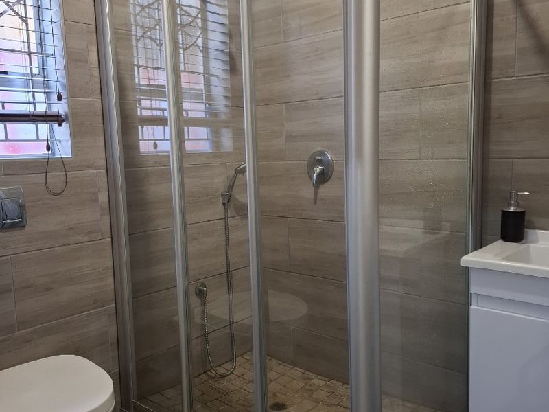 Modern bathroom featuring a glass shower enclosure, wall-mounted toilet, and a vanity cabinet with basin, finished with neutral wood-look wall and floor tiles.