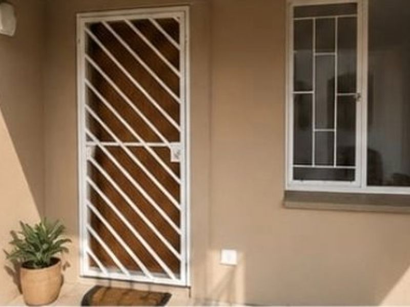 A clean apartment entrance featuring a white security gate and matching window security bars on a neutral-toned exterior wall.
