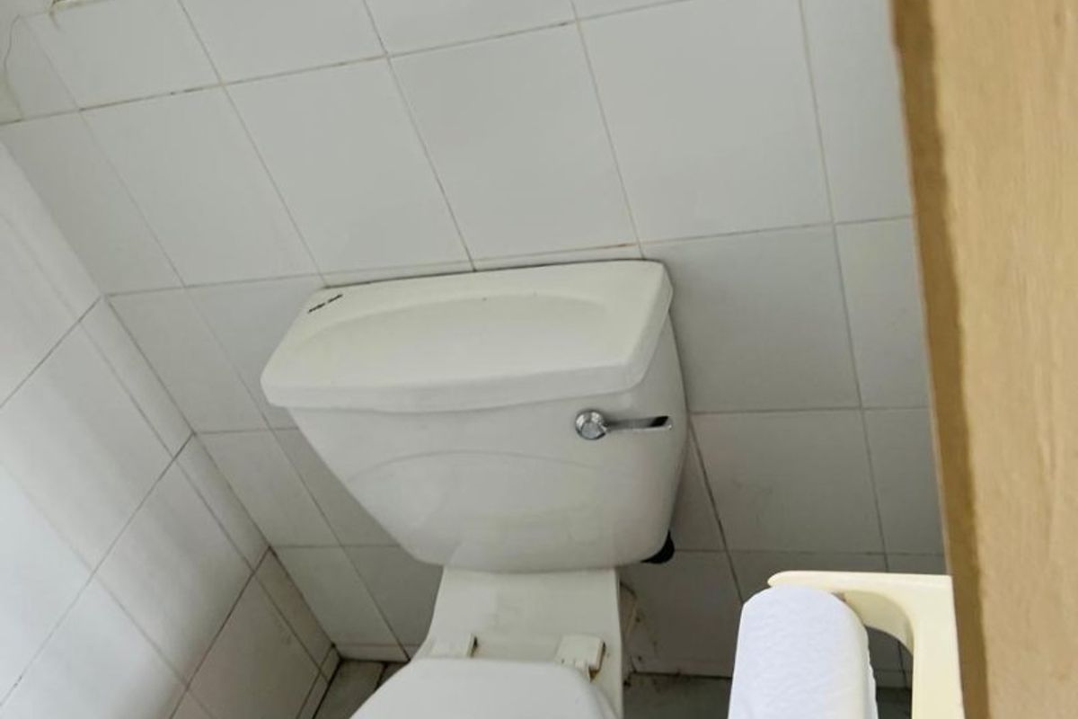 A compact bathroom featuring white wall tiles, a standard toilet, and a window with security bars.