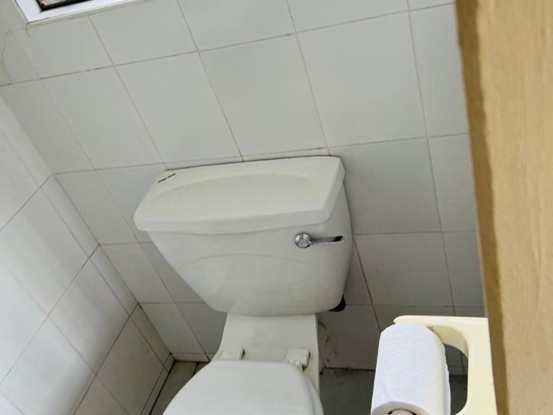 A compact bathroom featuring white wall tiles, a standard toilet, and a window with security bars.