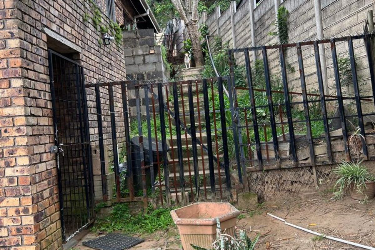 Rear exterior area featuring a brick building facade, concrete block boundary walls, rusted metal fencing, and concrete stairs leading to an elevated garden section.