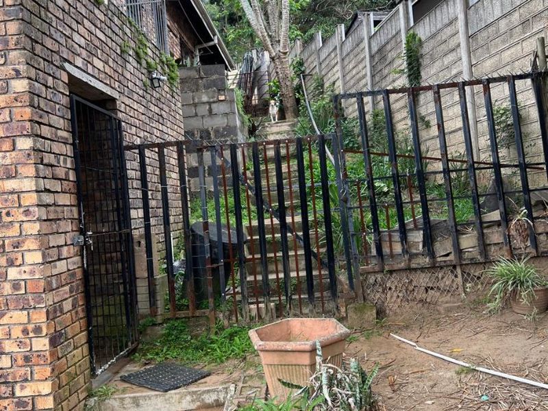 Rear exterior area featuring a brick building facade, concrete block boundary walls, rusted metal fencing, and concrete stairs leading to an elevated garden section.
