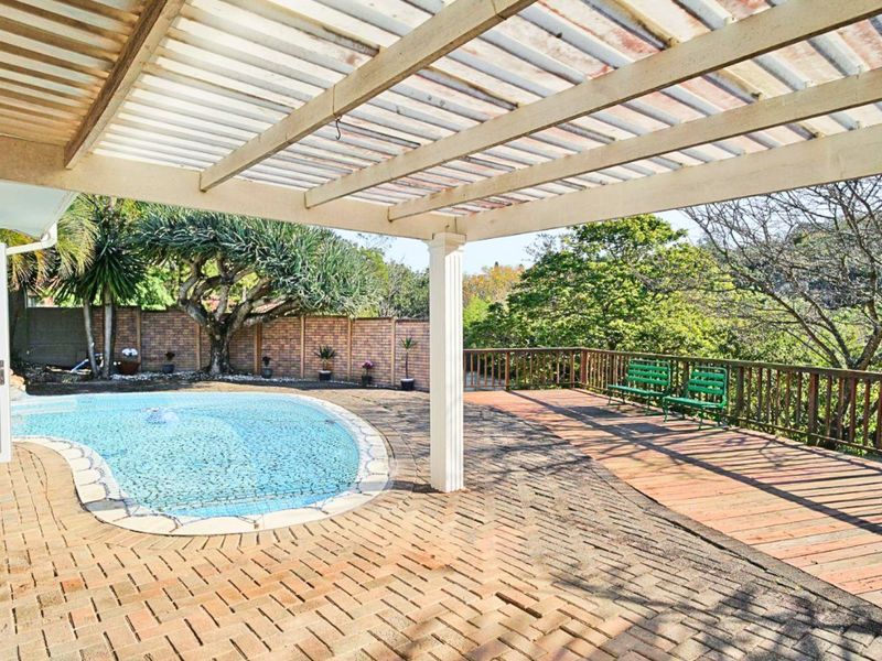 Outdoor entertainment area featuring a swimming pool, covered pergola, timber decking, and brick paving, overlooking a garden with a brick boundary wall.