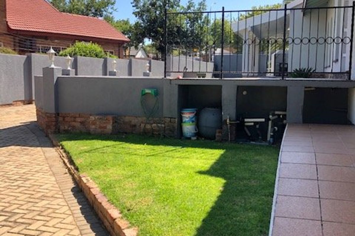 Rear exterior area featuring a paved driveway, landscaped lawn, brick retaining wall, and a dedicated housing area for pool filtration equipment.