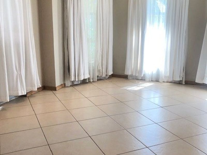 A bright, empty living area featuring neutral ceramic floor tiles and large windows with floor-to-ceiling curtains.