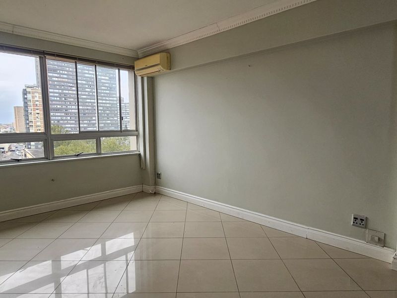 A bright, empty living room featuring large windows with city views, tiled flooring, and a wall-mounted air conditioning unit.