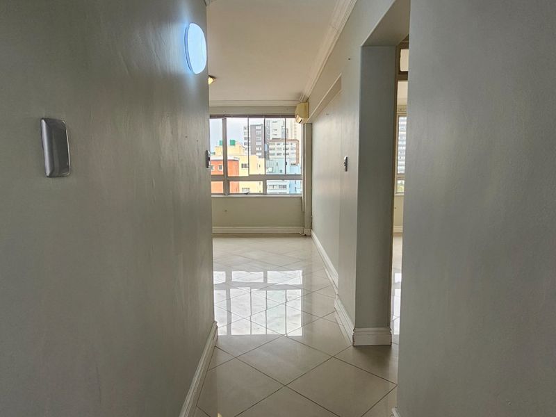 A bright, empty hallway with neutral walls and tiled flooring, leading towards an open living area with large windows.