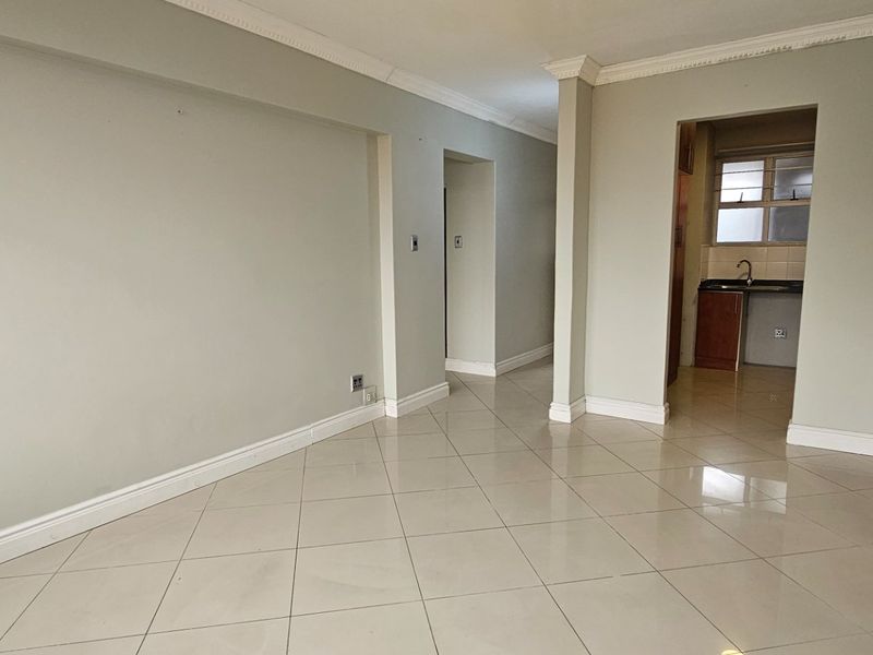 An empty, well-maintained living area featuring neutral walls, tiled flooring, and classic crown molding, with a view into the adjacent kitchen space.