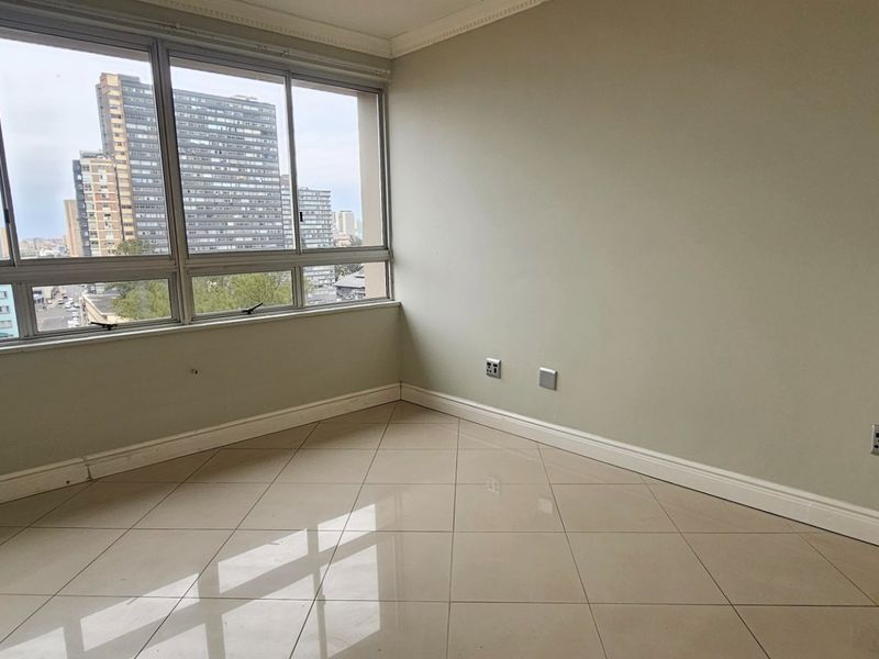 A bright, empty living area featuring large windows with city views, neutral walls, and polished tiled flooring.