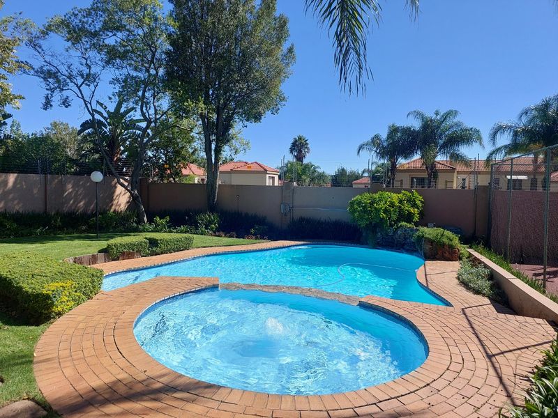 A well-maintained outdoor pool area featuring a curved swimming pool, brick paved surround, and established garden landscaping enclosed by a perimeter wall.