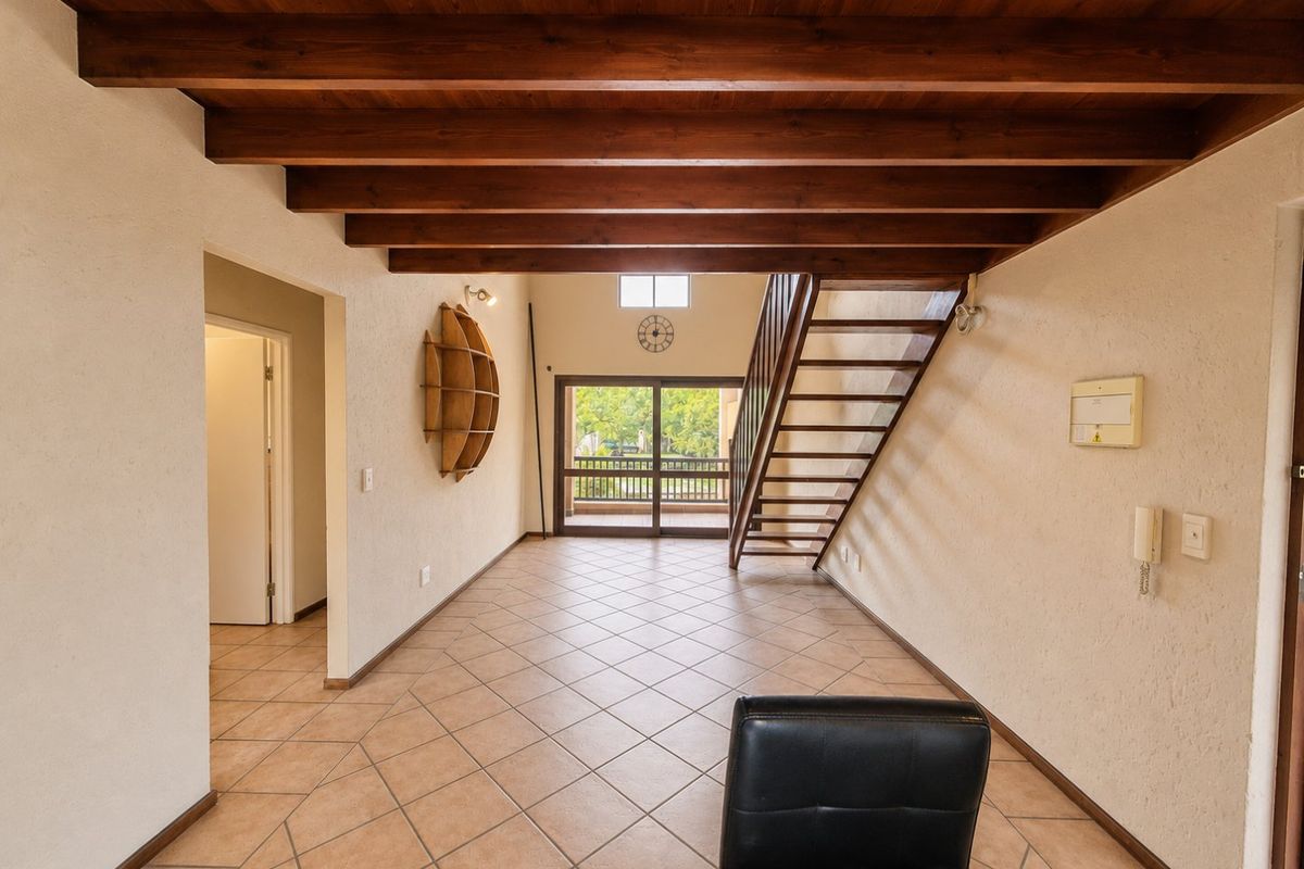 Spacious living area featuring exposed timber ceiling beams, tiled flooring, and a timber staircase leading to an upper level with balcony access.
