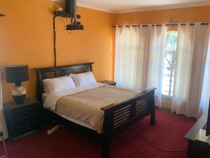 A bedroom featuring orange walls, red carpeting, and a ceiling-mounted projector. The room includes a window with security bars and standard curtain fixtures.
