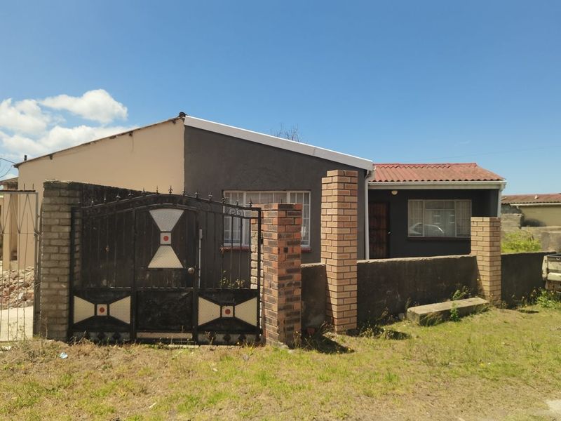 Front exterior view of a single-story house featuring a gated entrance, brick boundary wall, and corrugated metal roofing.