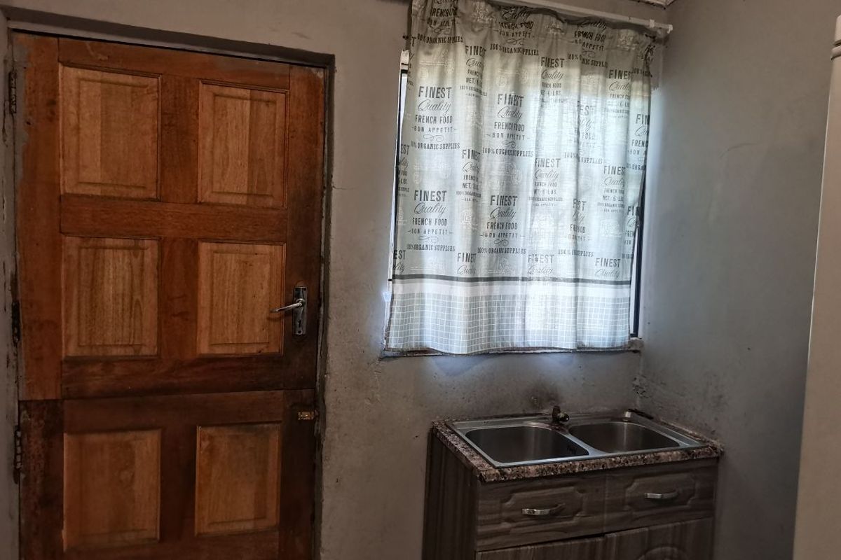 A small kitchen area featuring a double bowl stainless steel sink, built-in cabinetry, and a wooden door, showing signs of wear and wall deterioration.