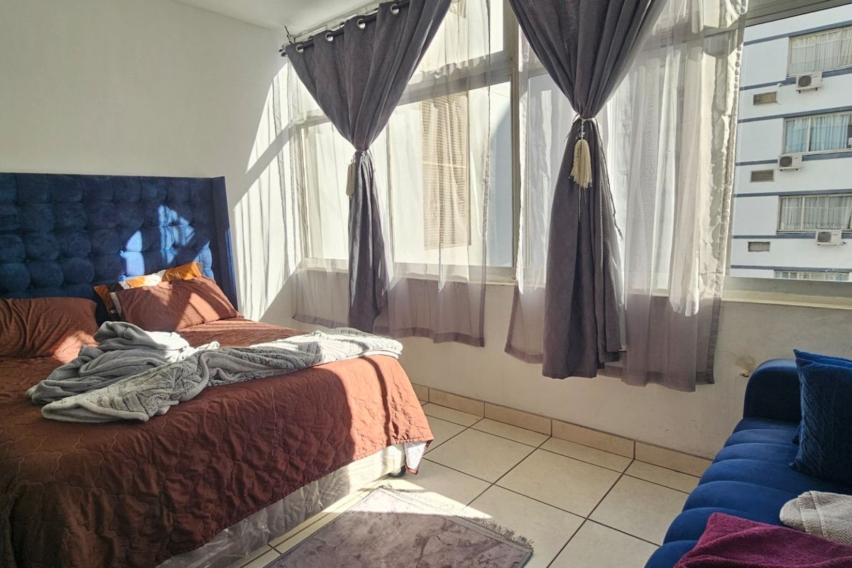 A sunlit bedroom featuring tiled flooring and large windows, offering a bright and functional living space.