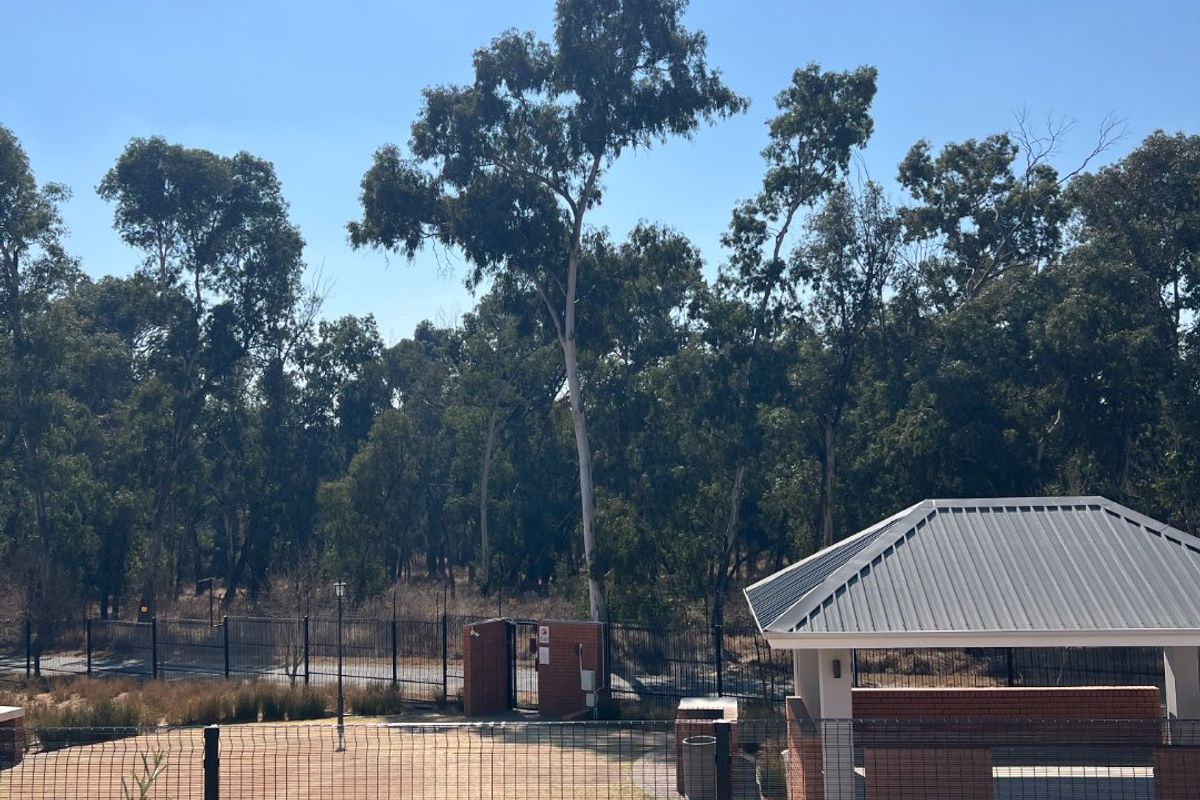 A well-maintained outdoor communal area featuring a brick gazebo with a metal roof, enclosed by wire mesh fencing and overlooking a wooded area.