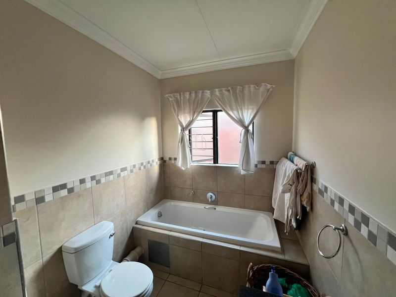 A functional bathroom featuring a built-in bathtub, toilet, and neutral tiled walls with a decorative border, complemented by a window with security bars.