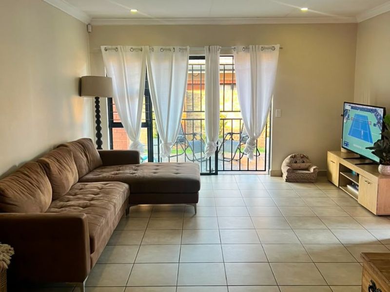 A spacious living room featuring neutral tiled flooring, recessed downlighting, and a sliding glass door providing access to an outdoor area with security gate.