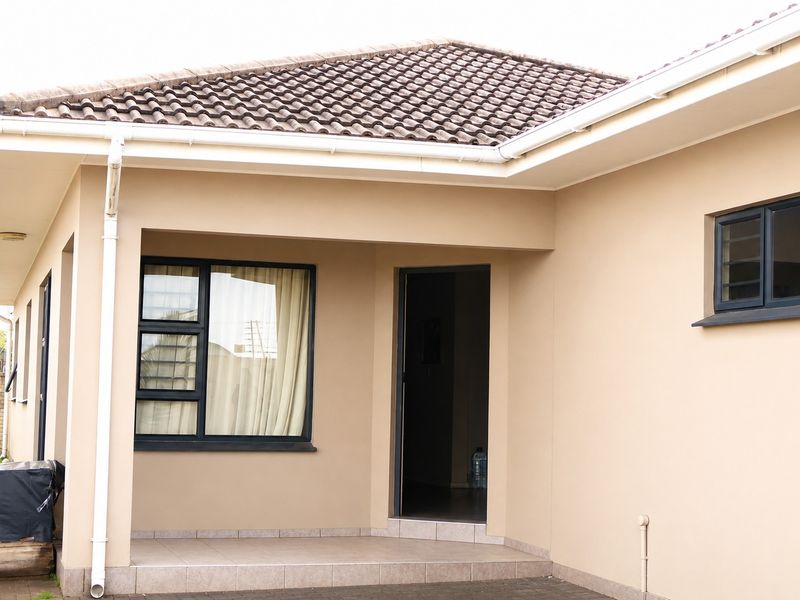 A well-maintained exterior view of the apartment entrance featuring a covered tiled patio, modern aluminium window frames, and a clean tiled roof.