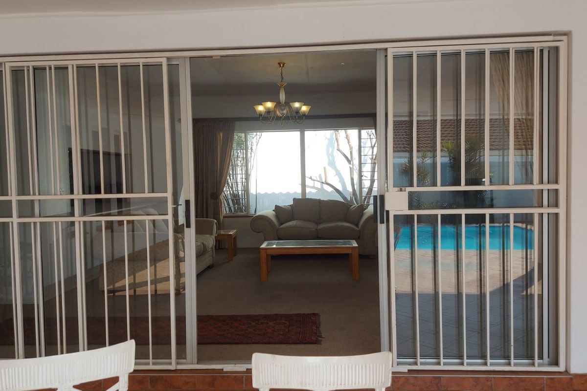 A living room viewed through sliding glass security doors, featuring large windows that provide natural light and a view of the outdoor pool area.