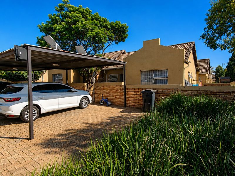 Front exterior view of the townhouse featuring a paved driveway, covered carport, and brick boundary wall with established garden landscaping.