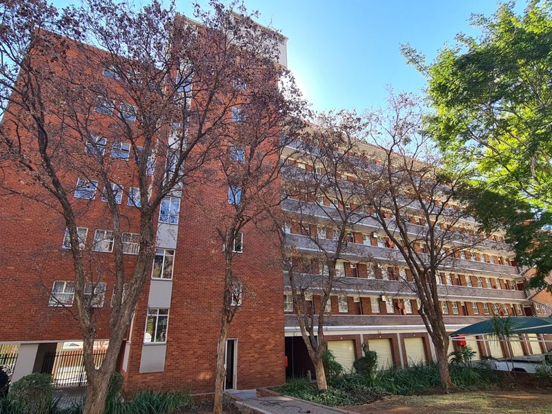 A multi-story commercial brick building featuring covered parking bays and established trees in the foreground.