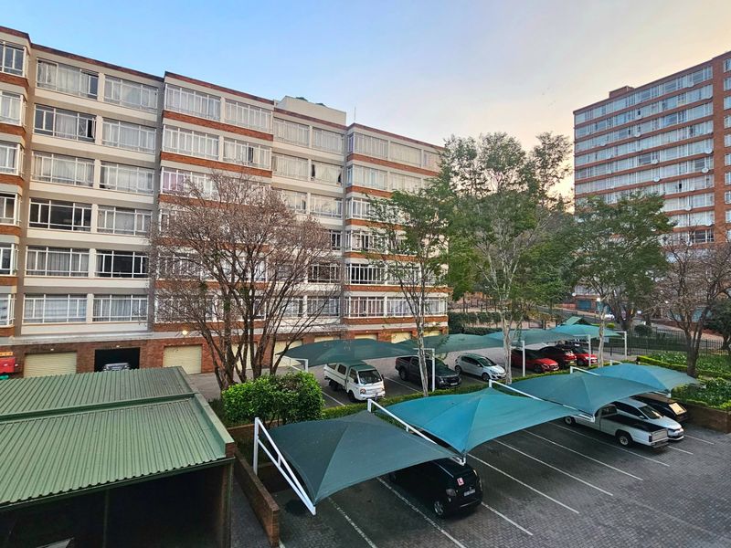 Exterior view of a multi-story brick apartment complex featuring paved parking areas with shade cloth structures and established landscaping.