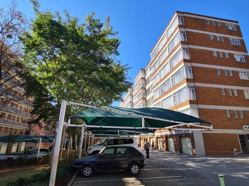 Exterior view of the Manitoba apartment complex featuring a multi-story brick facade, paved parking area, and covered shade cloth carports.