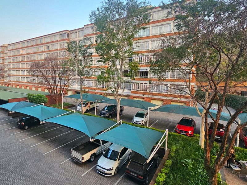 An elevated view of a commercial property featuring a large multi-story brick building with extensive window banks and a paved parking area with shade carports.