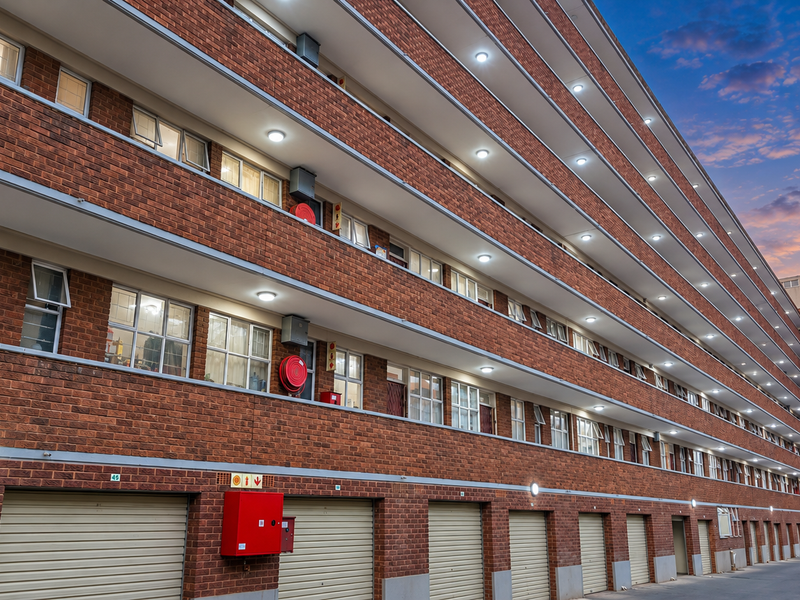 A multi-story commercial or residential building featuring a face brick facade, multiple ground-level garage units, and external corridor lighting at dusk.