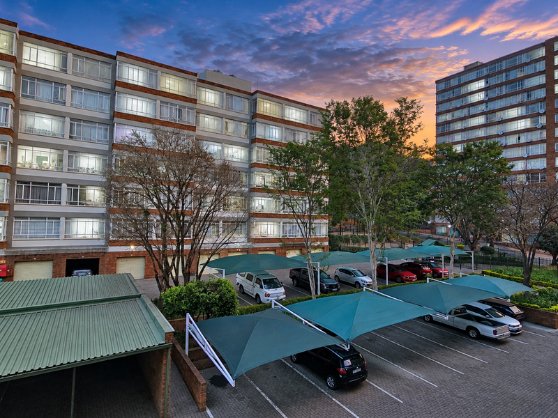 Exterior view of a multi-story commercial or residential complex featuring brick facades, paved parking areas, and shade cloth carports at sunset.
