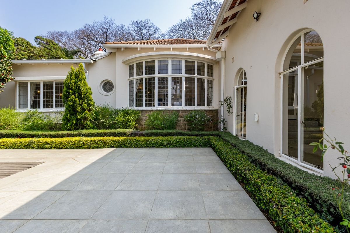 A well-maintained rear exterior view featuring a paved patio, manicured hedges, and classic architectural details including arched windows and a bay window.